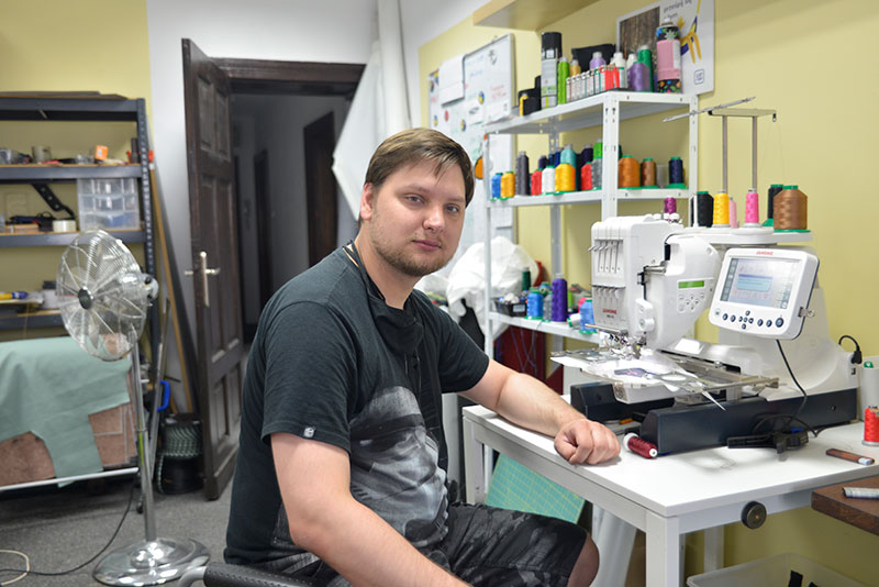 Fotografia przedstawia Jerzego Janiaka, młodego mężczyznę o krótkich włosach, który siedzi przy jednej z maszyn w zakładzie krawieckim. Autorem zdjęcia jest Dominik Wójcik.
