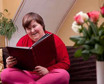 Fotografia przedstawia kobietę o krótkich włosach, w czerwonym swetrze, która siedzi po turecku na kanapie. Przegląda książkę, prawdopodobnie jest to jakiś album. Zdjęcie pochodzi z Obrazy licencjonowane przez Depositphotos.com/Drukarnia Chroma).