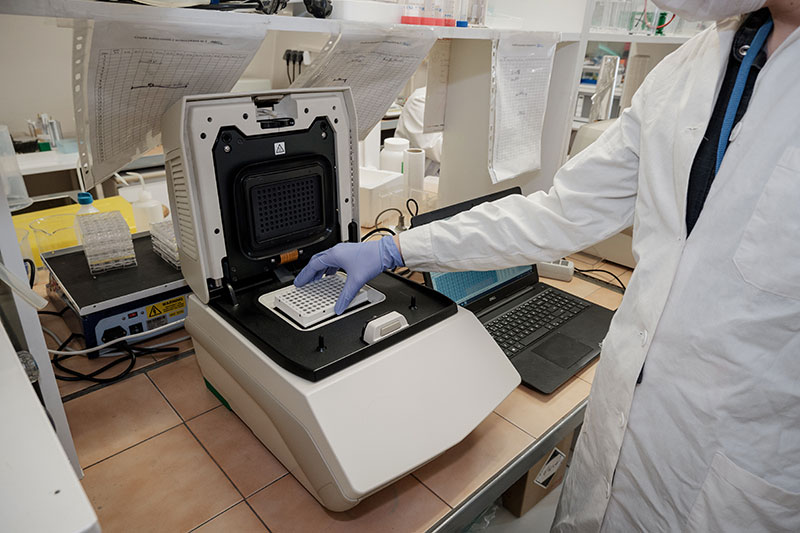 Na zdjęciu widoczna jest aparatura laboratoryjna obsługiwana przez laboranta w białym fartuchu i rękawiczkach. Laborant wkłada preparat do urządzenia. Autorem zdjęcia jest Konrad Kuczyński.