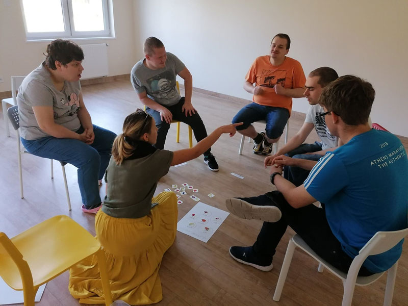Fotografia przedstawia sześć osób - pięcioro młodych osób z autyzmem i panią terapeutkę. Wszyscy siedzą na krzesłach, w kole i korzystają z zajęć. Zdjęcie pochodzi z materiałów Fundacji Dom Autysty.