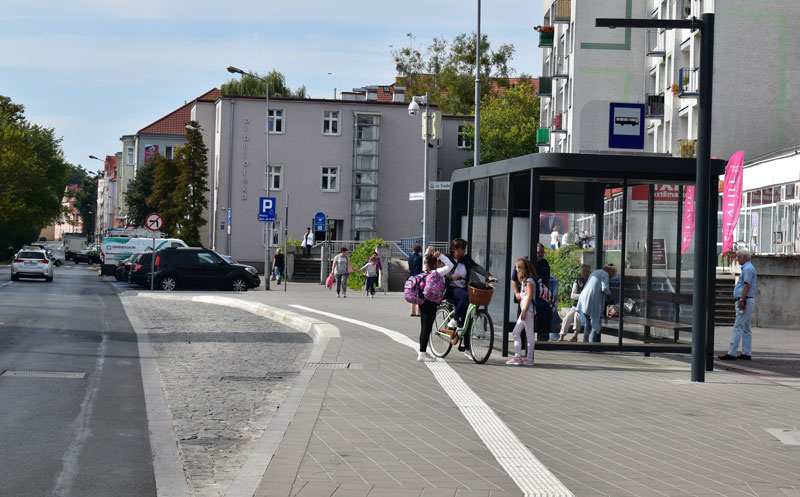 Fotografia przedstawia nowoczesny, czarny przystanek fotowoltaiczny przy jednej z ulic w Pile. Widocznych jest kilka osób – starszych i dzieci, a także pani na rowerze. Autorką zdjęcia jest Aleksandra Kramkowska.
