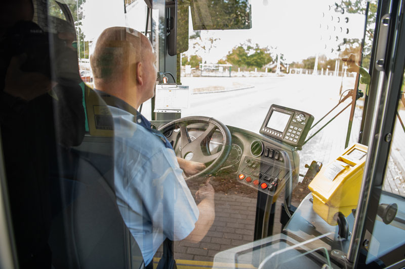 Fotografia przedstawia kierowcę autobusu, prowadzącego pojazd. Zdjęcie wykonane jest zza jego pleców. Autorem fotografii jest Maciej Motylewski.