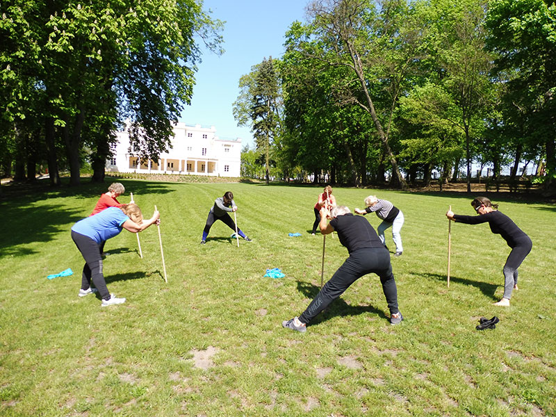 Fotografia przedstawia kilku seniorów, którzy w słonecznej pogodzie, na polanie przed pałacem, wykonują ćwiczenia gimnastyczne. Stoją w okręgu i opierają się, dwiema rękami, o kij. Zdjęcie pochodzi z archiwum beneficjenta.