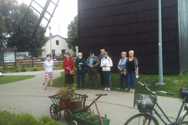 Fotografia przedstawia grupę seniorów pozujących na tle wysokiego, drewnianego wiatraka. Zdjęcie pochodzi z archiwum beneficjenta.