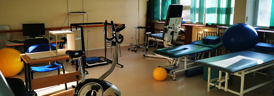 Fotografia przedstawia pomieszczenie przeznaczone do rehabilitacji ruchowej. Widoczne są między innymi rowerki stacjonarne, kozetki, większe i mniejsze gumowe piłki. Zdjęcie pochodzi z archiwum beneficjenta.