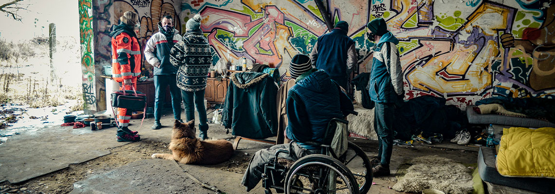 Fotografia przedstawia pustostan, w którym urządzili się bezdomni. Widać zniesione przez nich meble i przedmioty, a także liczne graffiti. Na zdjęciu widać sześć osób – pięcioro bezdomnych i ratowniczkę medyczną w pomarańczowym, odblaskowym stroju służbowym. Jeden z bezdomnych porusza się na wózku inwalidzkim. Przed nim leży duży pies. Autorem zdjęcia jest Dominik Wójcik.