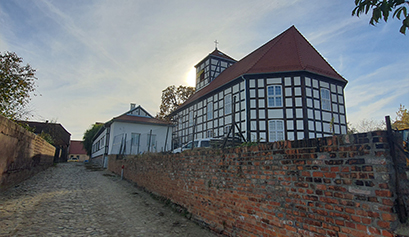 Fotografia przedstawia drogę ogrodzoną kamiennym murem, za którym znajduje się odrestaurowany kościółek ewangelicki. Zdjęcie pochodzi z archiwum beneficjenta.