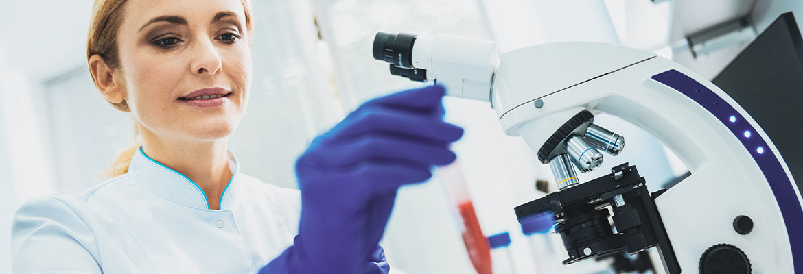 Fotografia przedstawia laborantkę w białym fartuchu i niebieskich rękawiczkach, pracującą przy mikroskopie. Zdjęcie pochodzi z Obrazy licencjonowane przez Depositphotos.com/Drukarnia Chroma.