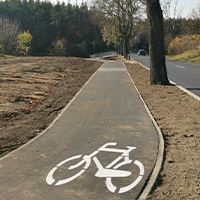 Fotografia przedstawia betonową ścieżkę rowerową biegnącą wzdłuż drogi. Zdjęcie pochodzi z archiwum beneficjenta.