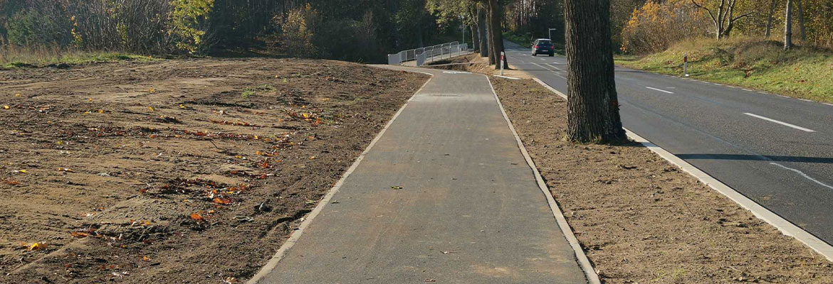 Fotografia przedstawia betonową ścieżkę rowerową biegnącą wzdłuż drogi. Zdjęcie pochodzi z archiwum beneficjenta.