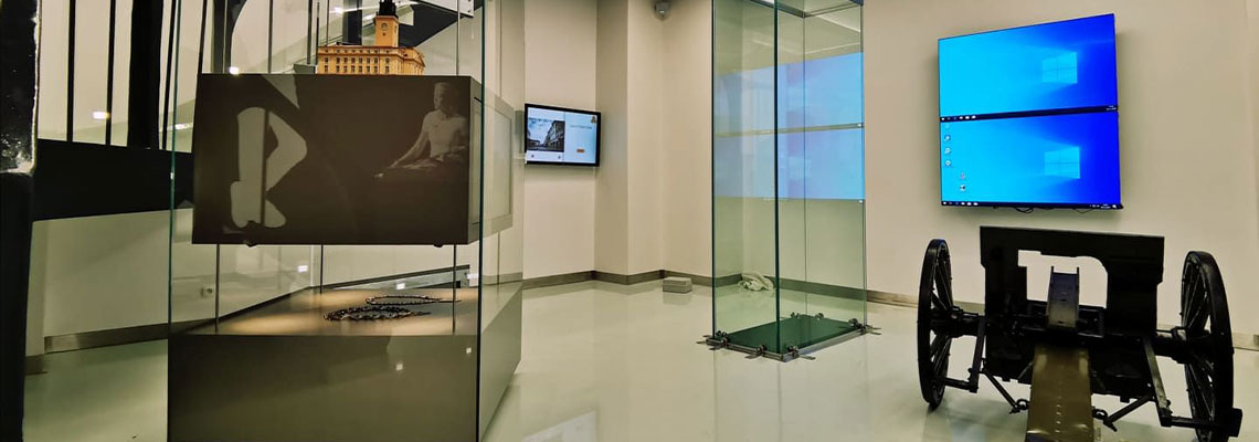 Fotografia przedstawia ekspozycję w kaliskim ratuszu. Widoczne są dwie przeszklone gabloty, monitory na ścianach oraz działo na kołach. Zdjęcie pochodzi z archiwum beneficjenta.