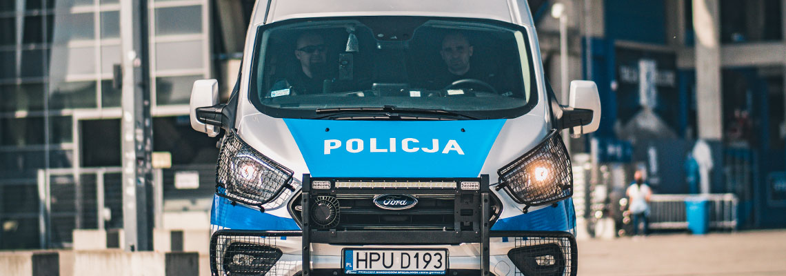 Fotografia przedstawia policyjny samochód od frontu. Ma tradycyjne – niebiesko-srebrne barwy i napis „Policja”. W środku widać dwóch funkcjonariuszy. Zdjęcie pochodzi z archiwum beneficjenta.