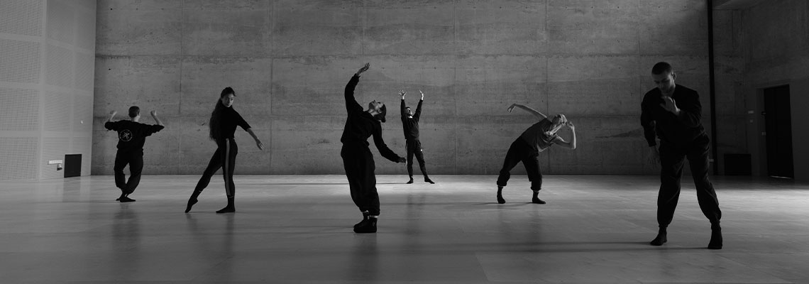 Czarno-biała fotografia przedstawia grupę sześciu tancerzy w różnych pozach, rozstawionych na parkiecie w przestronnej sali. Autorem zdjęcia jest Andrzej Grabowski.
