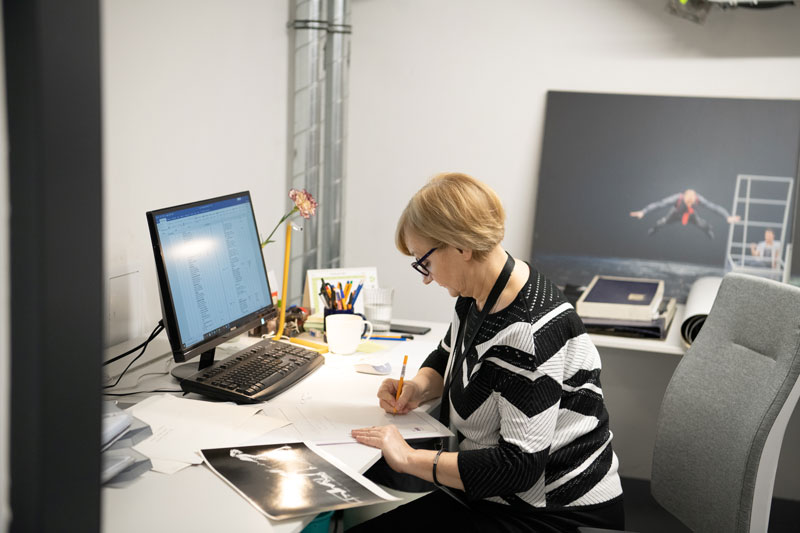 Fotografia przedstawia Małgorzatę Andrzejewską, archiwistkę Polskiego Teatru Tańca, w czasie pracy. Kobieta siedzi przy biurku, na którym znajduje się między innymi komputer, i w skupieniu wypełnia dokumenty. Autorem zdjęcia jest Maciej Motylewski.