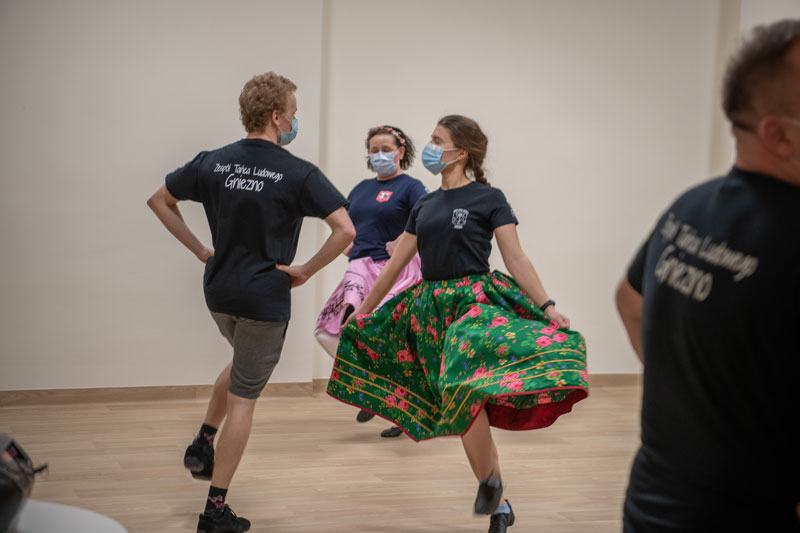 Fotografia przedstawia próbę Zespołu Tańca Ludowego „Gniezno”. Na głównym planie widać kobietę w rozłożystej spódnicy w kolorowe wzory oraz mężczyznę. Autorem zdjęcia jest Maciej Motylewski.