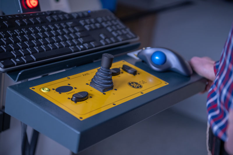 Fotografia przedstawia panel sterowniczy przemysłowego tomografu komputerowego. Pulpit jest żółty, widoczne są przyciski, joystick i myszka. Autorem zdjęcia jest Maciej Motylewski.
