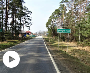 Fotografia przedstawia fragment drogi biegnącej przez las. Na dalszym planie znajduje się wjazd do miejscowości, o czym informuje znak po prawej stronie, z napisem „Szklarka Myśleniewska”. Zdjęcie pochodzi z archiwum WZDW w Poznaniu.