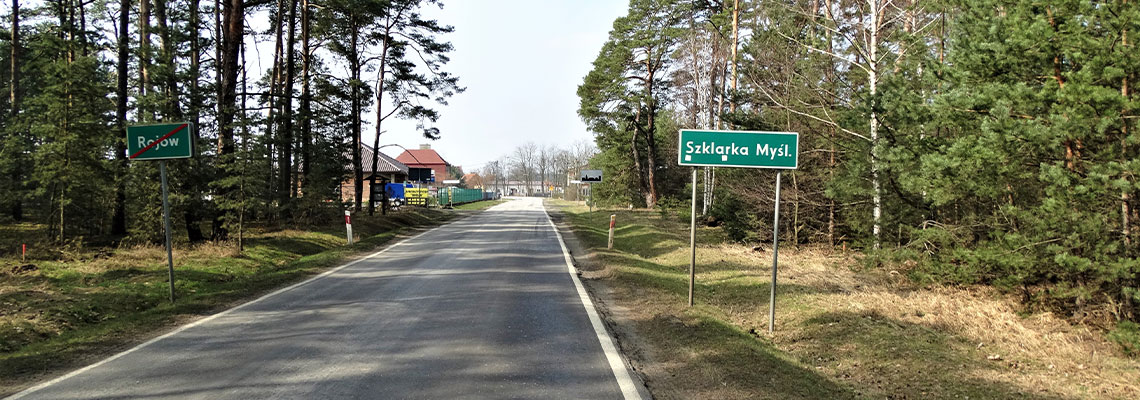 Fotografia przedstawia fragment drogi biegnącej przez las. Na dalszym planie znajduje się wjazd do miejscowości, o czym informuje znak po prawej stronie, z napisem „Szklarka Myśleniewska”. Zdjęcie pochodzi z archiwum WZDW w Poznaniu.