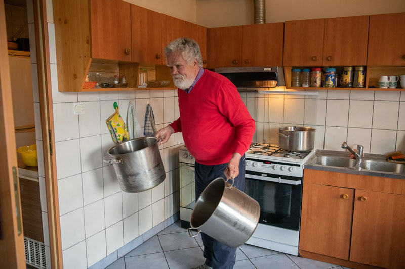 Fotografia przedstawia mężczyznę o siwych włosach i gęstej siwej brodzie, w czerwonym swetrze. Mężczyzna jest w kuchni, przenosi dwa wielkie srebrne garnki. Autorem zdjęcia jest Maciej Motylewski.