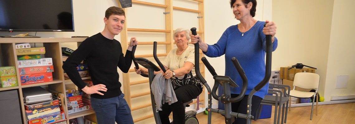 Fotografia przedstawia dwie starsze panie i młodego rehabilitanta. Jedna z pań korzysta z rowerka, druga ze steppera. Scena ma miejsce na sali gimnastycznej. Autorem zdjęcia jest Dominik Wójcik.