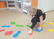 Fotografia przedstawia dziewczynkę, która porusza się po specjalnie do tego rozłożonych kolorowych znakach w kształcie ludzkich stóp, butów i dłoni. Zdjęcie pochodzi z archiwum beneficjenta.