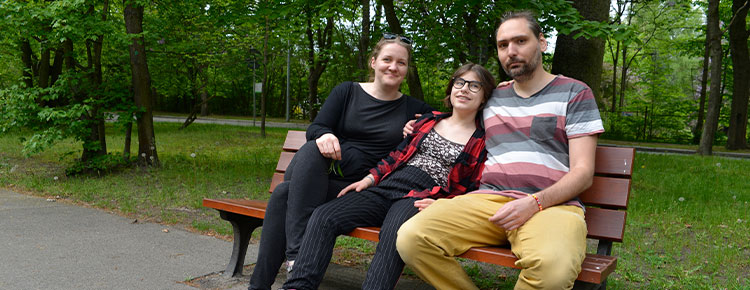 Fotografia przedstawia rodziców i córkę, siedzących na ławce w parku. Przytulają się. Po lewej siedzi matka, w środku córka na rolkach, po prawej ojciec w długich włosach. Autorem zdjęcia jest Dominik Wójcik.