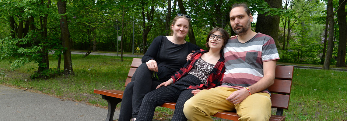Fotografia przedstawia rodziców i córkę, siedzących na ławce w parku. Przytulają się. Po lewej siedzi matka, w środku córka na rolkach, po prawej ojciec w długich włosach. Autorem zdjęcia jest Dominik Wójcik.