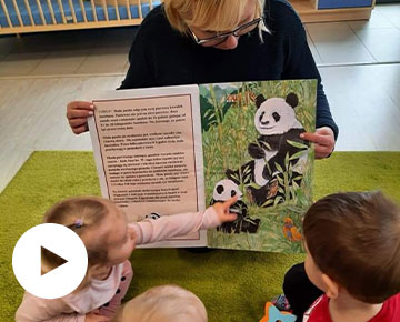 Fotografia przedstawia opiekunkę, siedzącą na dywanie przed trójką dzieci, trzymającą dużą książkę. Jedno z dzieci wskazuje na obrazek przedstawiający pandę. Zdjęcie pochodzi z archiwum beneficjenta.
