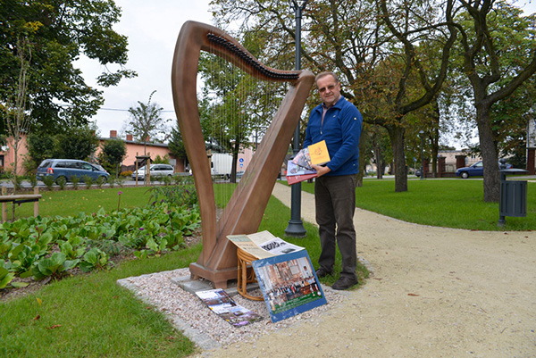 Fotografia przedstawia starszego mężczyznę w okularach, który trzyma w rękach kilka książek. Obok widoczna jest rzeźba harfy. Autorem zdjęcia jest Dominik Wójcik.