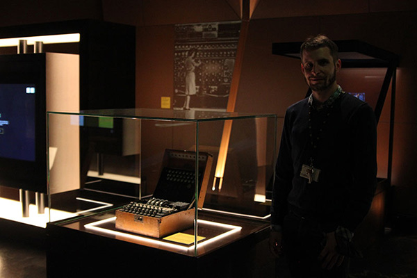 Fotografia przedstawia młodego mężczyznę o krótkich włosach i zaroście. Stoi obok szklanej gabloty, w której umieszczona jest Enigma. Autorem zdjęcia jest Dominik Wójcik.