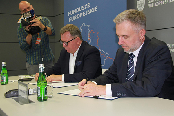 Fotografia przedstawia dwóch mężczyzn w garniturach, którzy siedząc przy stole, podpisują umowę. Obok stoi mężczyzna z kamerą. Zdjęcie pochodzi z archiwum Urzędu Marszałkowskiego Województwa Wielkopolskiego.