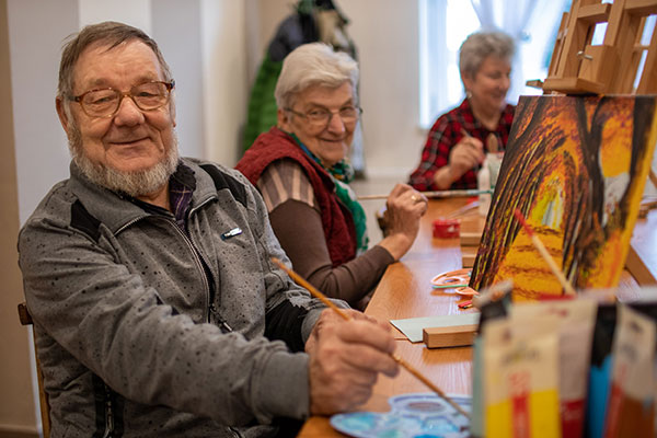 Fotografia przedstawia starszego, uśmiechniętego mężczyznę w okularach. Siedzi przy stole i maluje obraz ustawiony na sztaludze. Za nim siedzi starsza, uśmiechnięta kobieta, również malująca. W tle widać jeszcze jedną seniorkę. Autorem zdjęcia jest Bartłomiej Sroka.
