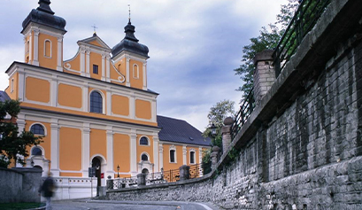Fotografia przedstawia fasadę kościoła. Widoczna jest droga, po prawej otoczona kamiennym murem. Sam kościół jest w jasnym kolorze, ma dwie wieże. Autorem zdjęcia jest Piotr Skórnicki.