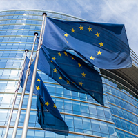 Fotografia przedstawia rząd unijnych flag – żółte gwiazdy ułożone w okrąg, na niebieskim tle, które powiewają przed przeszklonym gmachem. Zdjęcie pochodzi z Obrazy licencjonowane przez Depositphotos.com/Drukarnia Chroma.