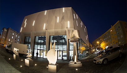 Nocna fotografia przedstawia fasadę nowoczesnego budynku, którego elewacja jest podświetlona. Przed gmachem widoczne są rzeźby, po prawej stronie zaparkowane samochody. Zdjęcie pochodzi z archiwum beneficjenta.