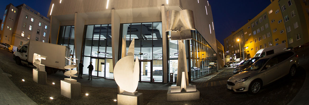 Nocna fotografia przedstawia fasadę nowoczesnego budynku, którego elewacja jest podświetlona. Przed gmachem widoczne są rzeźby, po prawej stronie zaparkowane samochody. Zdjęcie pochodzi z archiwum beneficjenta. 
