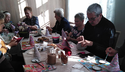 Fotografia przedstawia kilku seniorów, którzy przy stole tworzą ozdoby. Widoczne są również pierniki. Zdjęcie pochodzi z archiwum beneficjenta.