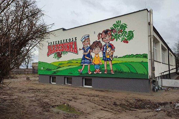 Fotografia przedstawia ścianę budynku – przedszkola. Znajduje się na niej kolorowy mural, przedstawiający trzy dziecięce postacie stojące wśród zielonych pól. Jest również napis „Przedszkole Jarzębinka). Wokół budynku widoczny jest teren placu budowy. Zdjęcie pochodzi z archiwum beneficjenta.