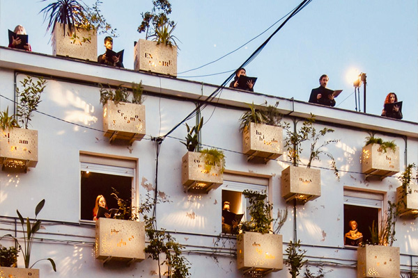 Fotografia przedstawia śpiewaków stojących na dachu i w oknach na najwyższej kondygnacji, w budynku, na którego elewacji zaczepiono zielone rośliny. Zdjęcie: Copyright: © European Union, 2021 .