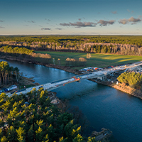 Fotografia z lotu ptaka przedstawia budowę mostu nad rzeką. Wokół widać gęsty las. Autorem zdjęcia jest Wojciech Waszak.
