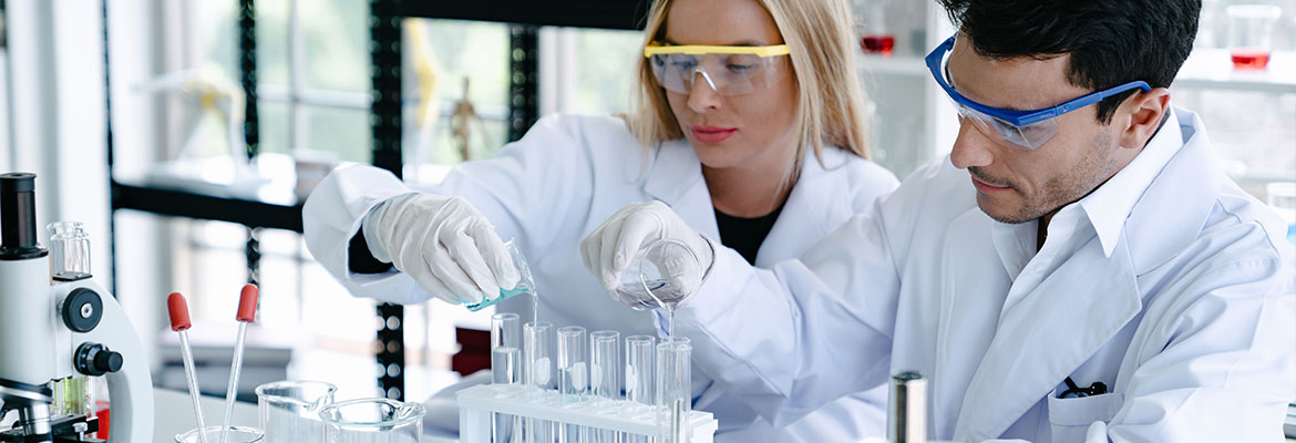 Fotografia przedstawia dwóch naukowców – kobietę i mężczyznę, w ochronnych okularach i białych fartuchach, pracujących w laboratorium. Zdjęcie pochodzi z adobestock.