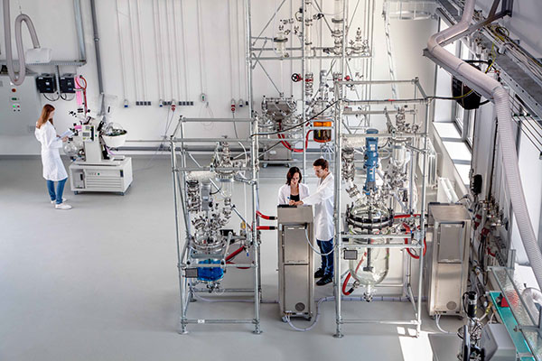 Fotografia przedstawia przestronne laboratorium ze skomplikowaną aparaturą badawczą. Przy urządzeniach stoją trzy osoby w białych fartuchach. Zdjęcie pochodzi z archiwum PPNT.