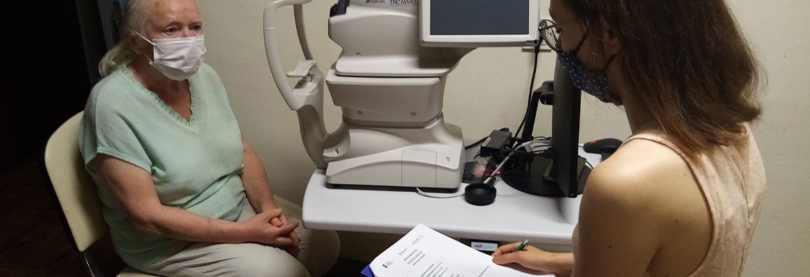 Fotografia przedstawia starszą kobietę siedzącą na krześle przed aparaturą do badania wzroku. Przed nią, tyłem do fotografującego, siedzi zaś osoba, która przeprowadza badanie. Zdjęcie pochodzi z archiwum beneficjenta.