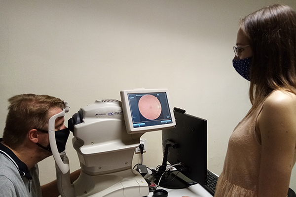 Fotografia przedstawia mężczyznę w trakcie badania wzroku. Spogląda w okular specjalnego urządzenia. Przed nim stoi kobieta, która wykonuje badanie. Zdjęcie pochodzi z archiwum beneficjenta.
