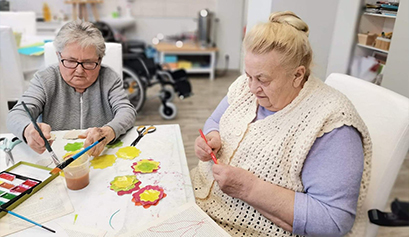 Fotografia przedstawia dwie starsze panie, które przy stole, w skupieniu, wykonują prace plastyczne. Zdjęcie pochodzi z archiwum beneficjenta.