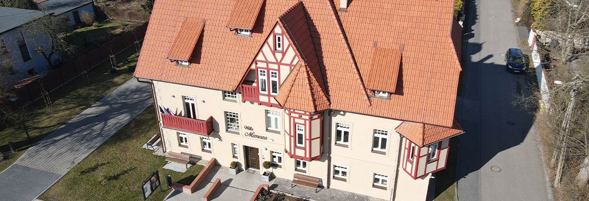 Fotografia z lotu ptaka przedstawia odnowiony budynek o nieregularnym kształcie z jasną elewacją i spadzistym dachem krytym czerwoną dachówką. Autorem zdjęcia jest Maciej Motylewski.