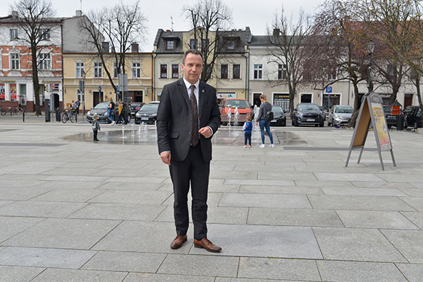 Fotografia przedstawia burmistrza Wojciecha Lisa. Mężczyzna w garniturze stoi na placu miejskim, w tle widać przechadzających się mieszkańców. Autorem zdjęcia jest Dominik Wójcik.