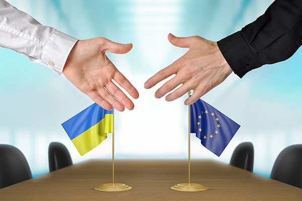 Fotografia przedstawia dwie dłonie osób, których nie widać, zmierzające do uścisku. W tle widoczny jest podłużny stół, na którym stoją flagi Ukrainy i Unii Europejskiej. Zdjęcie pochodzi z  Obrazy licencjonowane przez Depositphotos.com/Drukarnia Chroma.