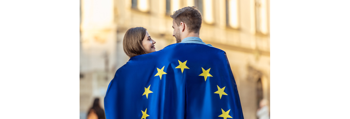 Fotografia przedstawia dwójkę młodych ludzi – dziewczynę i chłopaka, prawdopodobnie parę, którzy idą przed siebie, z flagą Unii Europejskiej narzuconą na plecach. Uśmiechają się. Zdjęcie pochodzi z Obrazy licencjonowane przez Depositphotos.com/Drukarnia Chroma