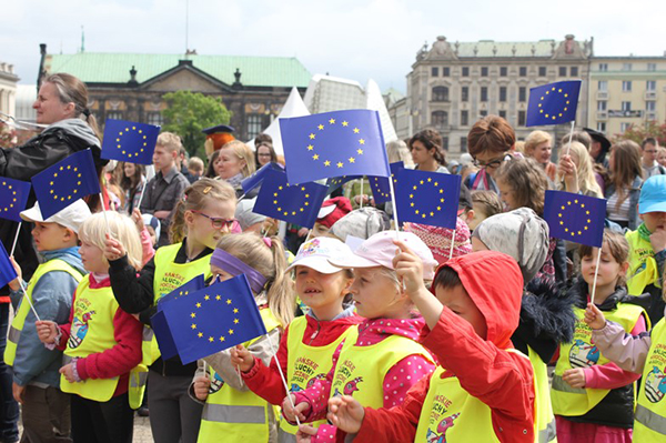 Fotografia przedstawia grupę przedszkolaków na imprezie plenerowej. Mają założone żółte kamizelki i machają wesoło unijnymi flagami. Zdjęcie pochodzi z archiwum Europe Direct Poznań.
